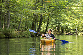 Kajak-Tourist paddelt auf einem Flussarm. Die Sonne strahlt durch den Wald, Biosphärenreservat, Schlepzig, Brandenburg, Deutschland