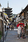 Young Japanese couple in kimono posing under a red umbrella in a narrow street with pagoda in the distance, Kyoto, Japan