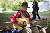 Folk musician with gray hair and a handlebar moustache playing a guitar while seated outdoors, a fiddle sitting beside him, United States of America