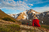 Hiker rests on a hillside overlooking Powerline Pass valley and trail, Chugach State Park, Southcentral Alaska