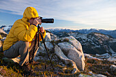A photographer uses a tripod and a zoom lens to capture a landscape photo from the top of a rocky mountain ridge.