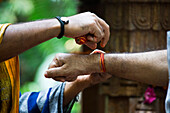 Nahaufnahme von einem Priester, der einem Gläubigen während einer Zeremonie einen Stofffaden ans Handgelenk bindet, Goa, Indien