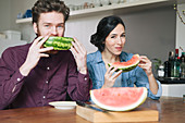 Portrait of happy young couple eating watermelon at kitchen table