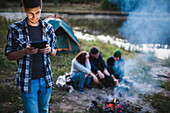 Young man using digital tablet while friends sitting by bonfire in forest
