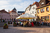 Menschen sitzen draußen vor Restaurants und lauschen der Musikkapelle Eußenhausen während einem kostenlosen Freitagabendskonzert am Marktplatz in der Altstadt, Bischofsheim, Rhön, Bayern, Deutschland