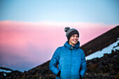 A woman in teal parka coat and wool hat stands in a sunset mountain scene.