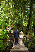 A young married couple hold hands while walking across a boardwalk in the forest.
