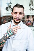 Barber store Schorem, Rotterdam, Netherlands. Portrait of a barber with tattoos on his arm, smoking a cigarette just outside the front door.
