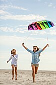 Two sisters run on the beach with a colorful kite in the sky above them.