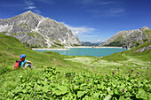 Woman hiking sitting in meadow and looking towards lake Luenersee, lake Luenersee, Raetikon trail, Raetikon, Vorarlberg, Austria