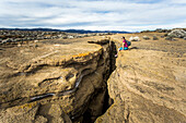 A young woman sits at the edge of a narrow, deep slot canyon in a remote desert