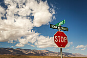 A stop sign and two street signs in the bottom corner of image, with desert mountains on horizon and a big, blue sky with puffy clouds