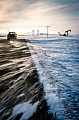 'A truck drives down a road with blowing snow and a pump jack in a snowy field at sunrise; Saskatchewan, Canada '