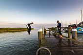 Three young people have fun and jump with a backflip from a pier in the Bodden by Ahrenshoop. Ahrenshoop, Althagen, Darß, Mecklenburg-Vorpommern, Germany