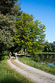 Two young women ride bicycles on path along Aubachseen lakes, Habichsthal, Spessart-Mainland, Bavaria, Germany