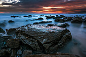 Water surrounds the rocks on the beach and the clouds glow orange and pink at sunset over the horizon, Tarifa, Cadiz, Andalusia, Spain