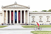 young woman bicycling in front of Glyptothek on Königs Plaza in Munich, Bavaria, Germany