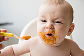 Mother feeding messy baby son with spoon in high chair