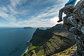 Mann sitzt am Rande einer Klippe auf einem Berg am Meer, Färöer Inseln