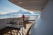 Ein Passagier steht an Deck von Expeditions Kreuzfahrtschiff MV Sea Spirit (Poseidon Expeditions) mit Blick auf schneebedeckte Berge, Gerlache Strait, Grahamland, Antarktische Halbinsel, Antarktis