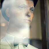 Old mannequin in shop window with hat and bow tie, reflections on glass