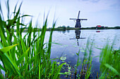 Green grass frames the windmill reflected in the canal at dusk Kinderdijk Rotterdam South Holland Netherlands Europe.