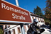 Motorcycle rider behind road sign for Romantische Strasse romantic road through Unteres Taubertal, Bronnbach, near Wertheim, Spessart-Mainland, Franconia, Baden-Wuerttemberg, Germany