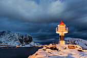 A lighthouse in the snow in the Arctic night with the village of Reine in the background Nordland Lofoten Islands Norway Europe
