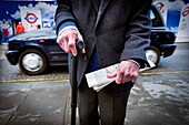 Primer plano de un hombre mayor irreconocible con un baston en una mano y un periodico en la otra, al fondo un taxi. Oxford St., Londres, Uk, Europa.