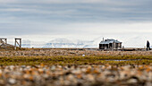 Man Walking Towards An Isolated Wooden House With Snowy Mountains In The Background