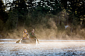 A Young Couple Paddles A Canoe Through Morning Mist On Long Pond Near Greenville, Maine