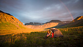 A Woman Is Sitting By Her Tent As A Rainbow Arcs Over Camp In Lake Clark National Park And Preserve