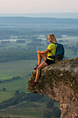 Woman sitting on sandstone overhang watching sunrise from summit of Petit Jean Mountain above Arkansas River Valley 