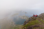 Hikers on top of rocky peak admire the blue sea surrounded by mist Reinebringen Moskenes Lofoten Islands Norway Europe