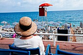 Woman with hat sitting on chair while loocking over 'Neptune Page' stone beach Nice, French Riviera, France