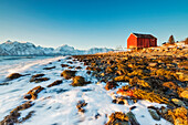 Typische Holzhütte namens Rorbu umgeben von Wellen des kalten Meeres und schneebedeckten Gipfeln, Djupvik, Lyngen Alpen, Troms, Norwegen, Skandinavien, Europa