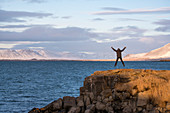 Eine Person springt in die Luft wie ein X vor verschneiter Landschaft an der Küste nahe Reykjavik mit Blick auf das Gebirge Akrafjall in Wolken, Reykjavik, Island, Iceland, Europa