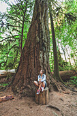 A young woman sits on a stump below an ancient Douglas Fir tree in Cathedral Grove, Cathedral Grove, British Columbia.