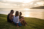Man and two women sitting on grass in front of ocean coastline, Kuta, Lombok, Indonesia