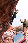 An athletic male boulders on a tall rock as another male spots him from below in Red Rocks, Nevada