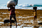 Surfer reiten Welle im Hintergrund und Menschen tragen Säcke im Vordergrund, Lakey Peak, Zentral-Sumbawa, Indonesien