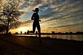 Silhouette of woman jogging on riverside road at cloudy sunset, Boston, Massachusetts, USA