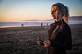 Junge Frau in der Jacke mit dem Pferdeschwanz, der Sonnenbrille auf Strand bei Sonnenuntergang, San Francisco, Kalifornien, USA hält.