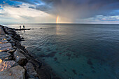 Rainbow and cloudy sky over silhouettes of two people fishing on coastal rocks, Massachusetts, USA