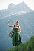 Junge Frau in Tracht macht ein Selfie mit ihrem Handy auf dem Falkenstein im Allgäu, Pfronten, Bayern, Deutschland