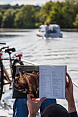 Hände von Mann beim Betrachten von auf Flohmarkt erworbenen Paul Bocuse-Kochbuch an Deck von Le Boat Elegance-Hausboot während Bootstörn auf Fluss Petit Saône, nahe Gray, Haute-Saône, Bourgogne Franche-Comté (Burgund), Frankreich, Europa