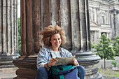 Portrait of smiling young Afro female tourist sitting with map against column at Altes Museum
