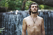 Shirtless young man with closed eyes standing against waterfall at forest