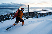 Kayak surfer carrying a paddle and kayak in winter along the snow covered shore, Homer, Southcentral Alaska, USA