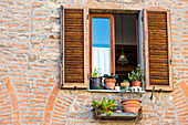 Close View Of A Montepulciano Brick House Facade With Opened Window, Flowerpots And Latticed Sun Blinds; Tuscany, Italy